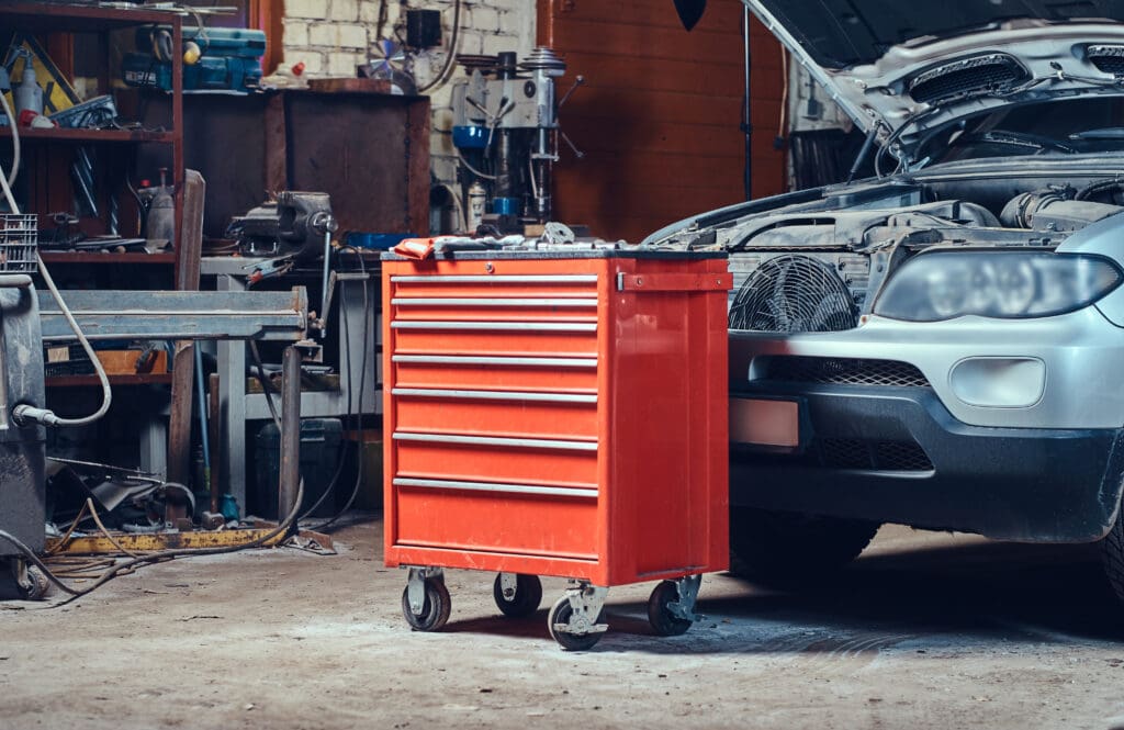 A red tool chest on wheels stands in a garage near a silver car with its hood open, ready for MOT testing. The garage is filled with various tools and equipment, creating a busy workshop atmosphere.