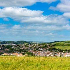 A scenic view of a lush green landscape with a small town nestled among the hills, where the sky is partly cloudy. Several houses and a local garage specializing in clutches are visible. A field with green grass is in the foreground, and rolling hills stretch into the distance.