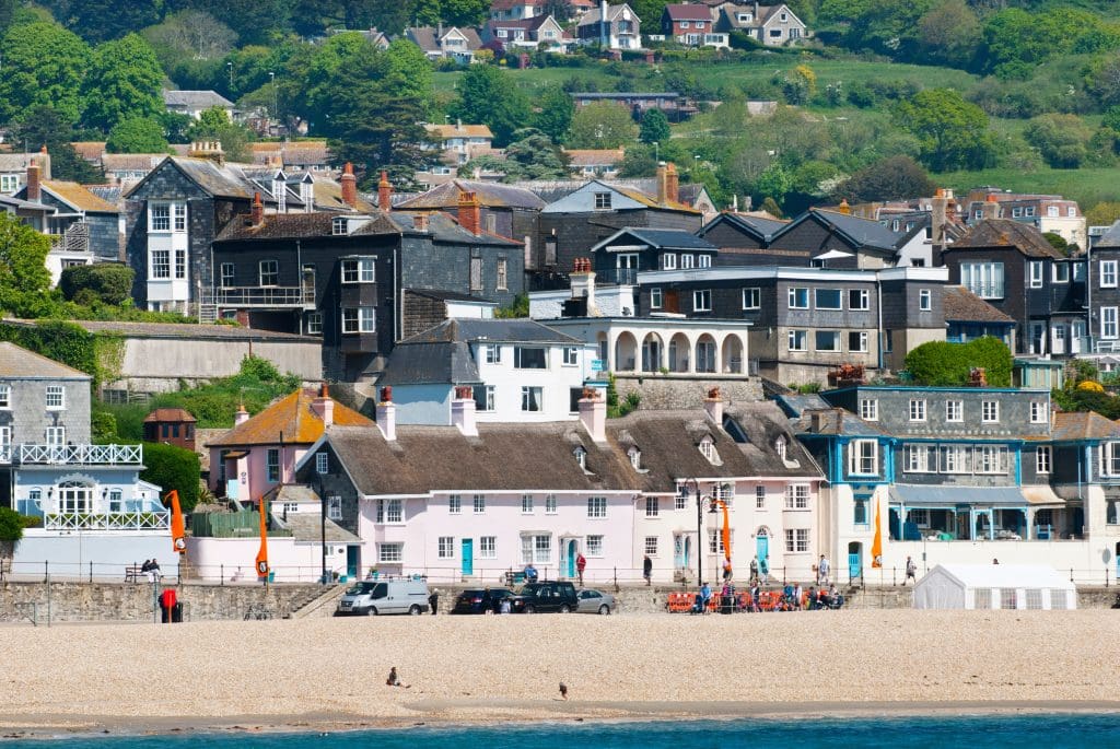 A coastal village with a sandy beach in the foreground, where colorful houses of varying architectural styles line the shore like neatly arranged clutches. Lush greenery and more homes are nestled on the hillside in the background.