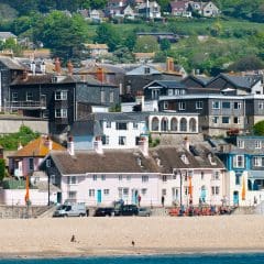 A coastal village with a sandy beach in the foreground, where colorful houses of varying architectural styles line the shore like neatly arranged clutches. Lush greenery and more homes are nestled on the hillside in the background.