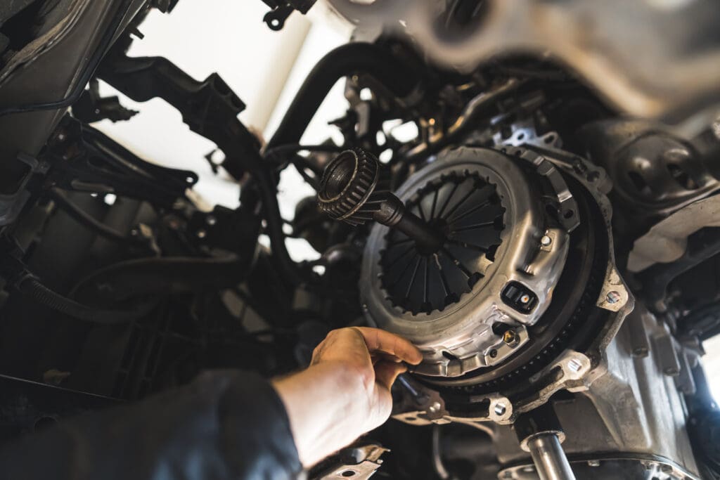 A garage mechanic's hand reaches towards a car's clutch assembly, part of the transmission system. The view from underneath the vehicle reveals various mechanical components, highlighting the thoroughness typical of car servicing or MOT testing.