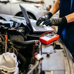 A mechanic wearing black gloves works on a car engine, using a laptop placed on the open hood. A digital multimeter is nearby, connected to the engine. Another car is visible in the background.