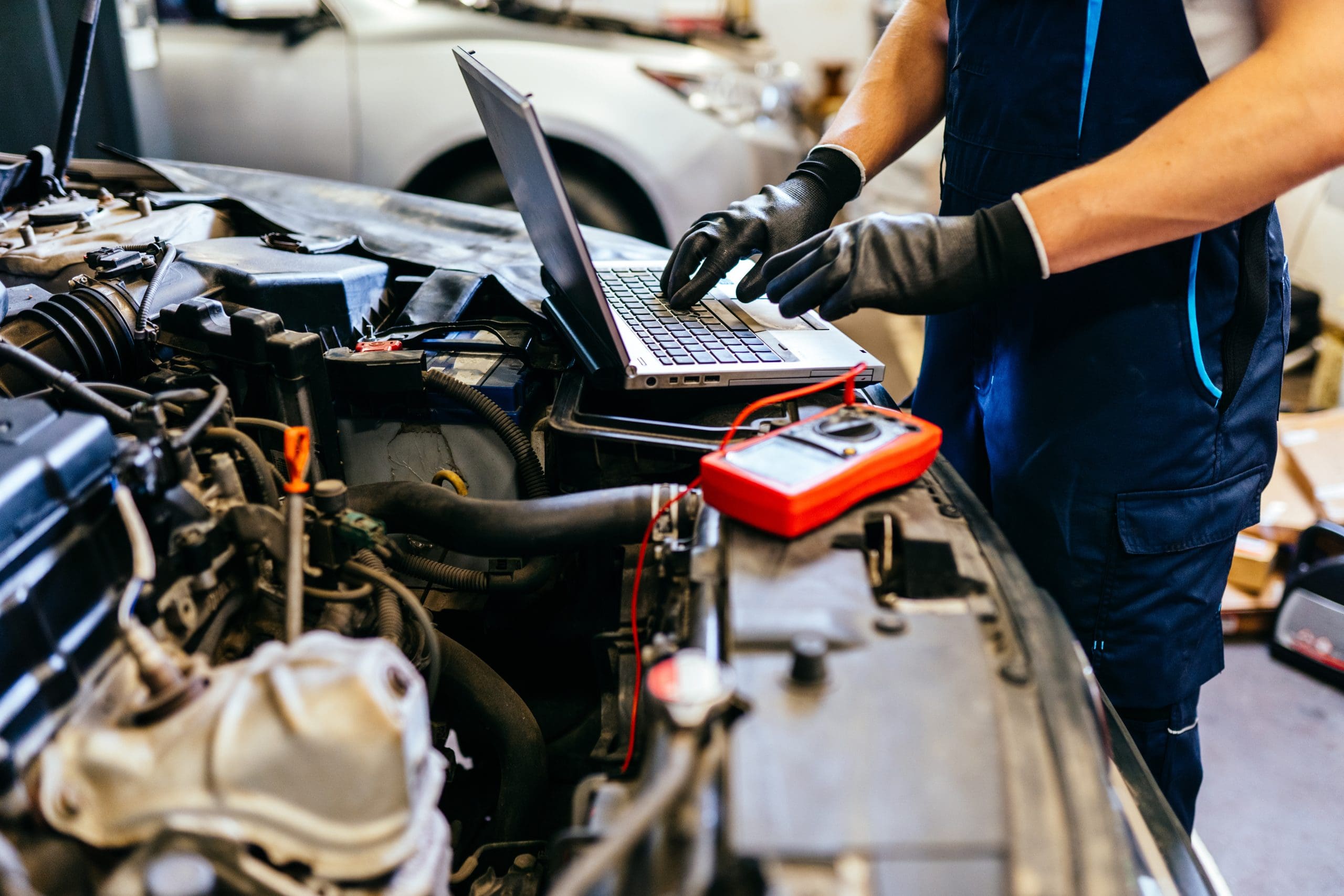 A mechanic wearing black gloves works on a car engine, using a laptop placed on the open hood. A digital multimeter is nearby, connected to the engine. Another car is visible in the background.