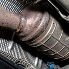 Close-up view of a car’s catalytic converter attached to the exhaust pipe, showing a metallic, cylindrical component with some rust and protective shielding in the surrounding area.