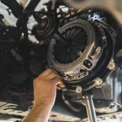 A mechanic’s hand works on a car’s clutch and transmission system from underneath the vehicle, adjusting components with tools. The view is close-up, showing gears and metal parts in detail.