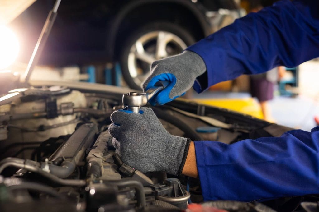 A mechanic wearing blue gloves and a blue uniform works on a car engine, using a socket wrench to tighten or loosen a part under the hood in a well-lit auto repair shop.