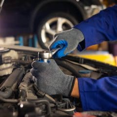 A mechanic wearing blue gloves and a blue uniform works on a car engine, using a socket wrench to tighten or loosen a part under the hood in a well-lit auto repair shop.